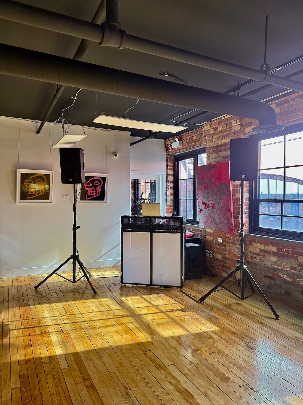 Two speakers on stands flanking a DJ booth and mixer in a sunlit loft space with exposed brick
